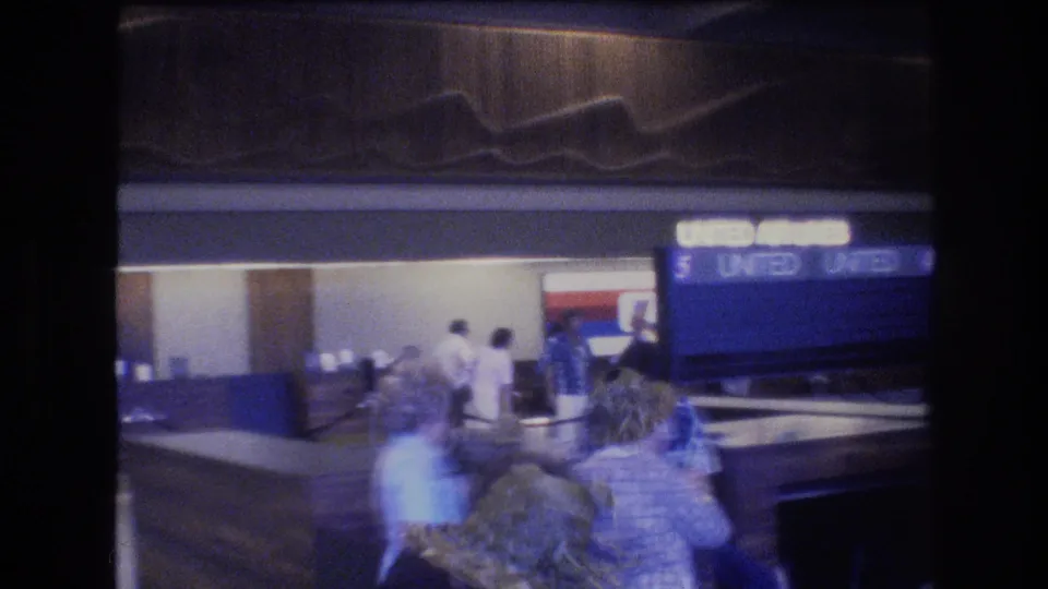 A United Airlines terminal counter appears behind passing travelers in 1977 Honolulu airport footage, with blue signage and blurred foreground movement.