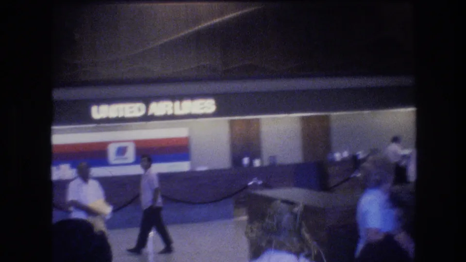 United Airlines signage is visible above an airport counter as travelers walk through the Honolulu terminal in soft, grainy 8mm footage from 1977.