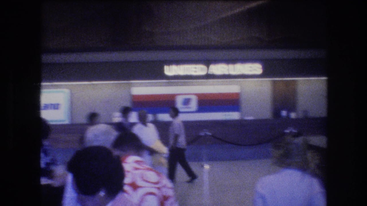 1977 Honolulu airport home-movie frame showing travelers near a United Airlines check-in counter with a red, white and blue logo panel.