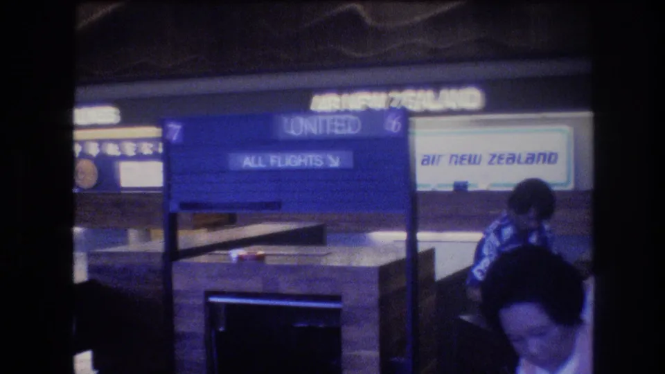 1977 Honolulu airport terminal view with United and Air New Zealand signage, an information board, and passengers partly visible in the foreground.