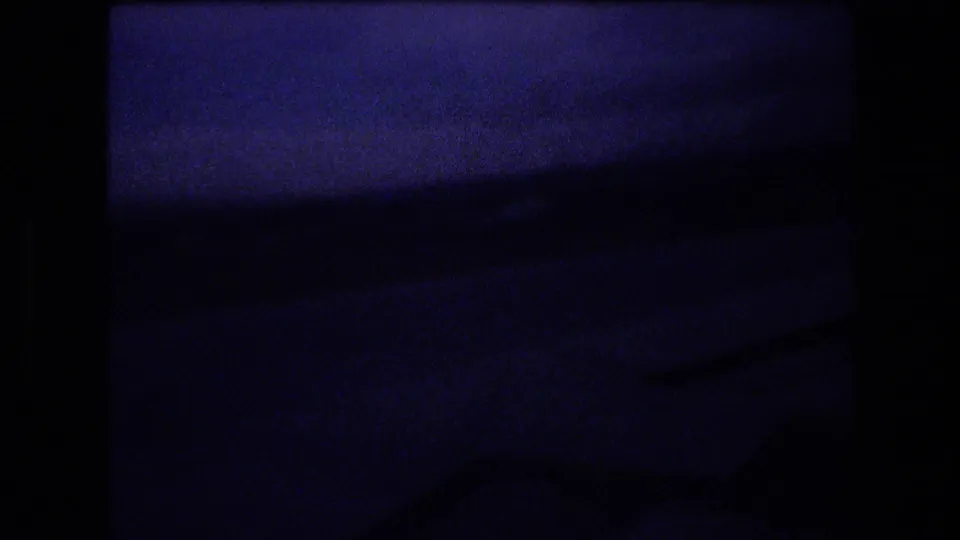 Blue-shifted, underexposed 8mm frame from the airport sequence, showing a dark interior or window area with minimal readable detail.