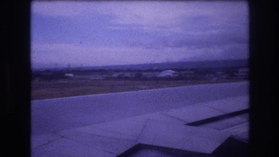 1977 Honolulu airport airplane-window view showing the wing over runway pavement and distant airport grounds under an overcast sky.