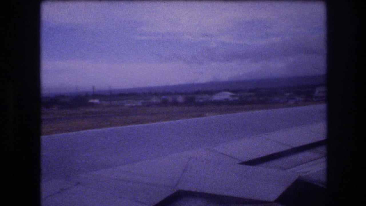1977 8mm passenger-window view likely over Oahu, Hawaii, with an airplane wing crossing the foreground and a hazy shoreline below.