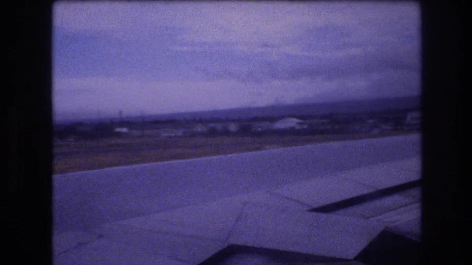 1977 8mm passenger-window view likely over Oahu, Hawaii, with an airplane wing crossing the foreground and a hazy shoreline below.