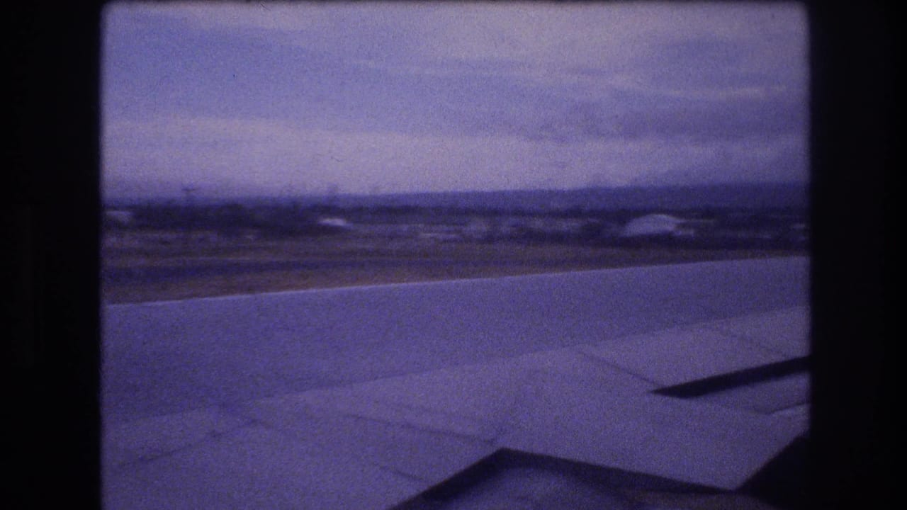 Airplane wing foreground with blurred coastal landscape and overcast sky beyond.