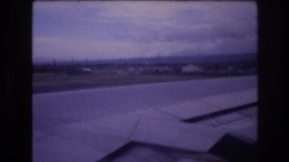 Vintage 8mm frame showing a passenger-seat view over likely Oahu coastal terrain, with the aircraft wing low in the frame and heavy film grain.