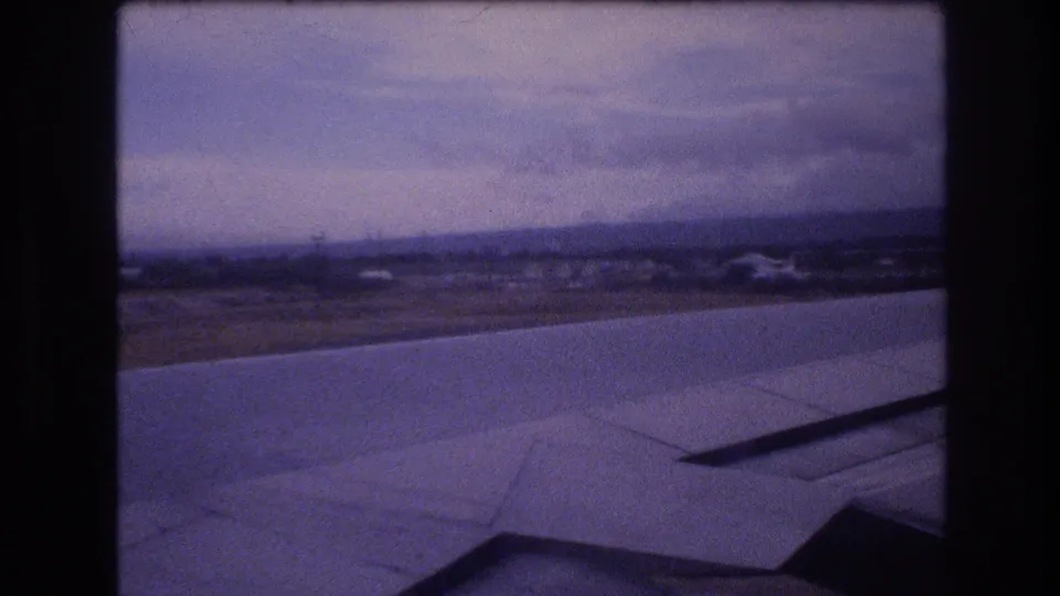 1977 home-movie frame likely filmed over Oahu, Hawaii, showing the airplane wing, distant coastal terrain, and purple-shifted sky through the aircraft window.