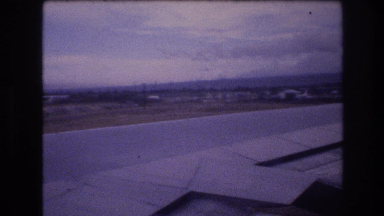 1977 Honolulu-area home-movie frame looking past an airplane wing toward runway pavement, distant airport structures, and an overcast horizon.