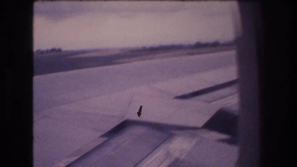 Soft, motion-blurred 8mm frame from an airplane window, showing the wing, runway surface, low horizon, and dark window-edge vignetting during takeoff or landing in Honolulu.