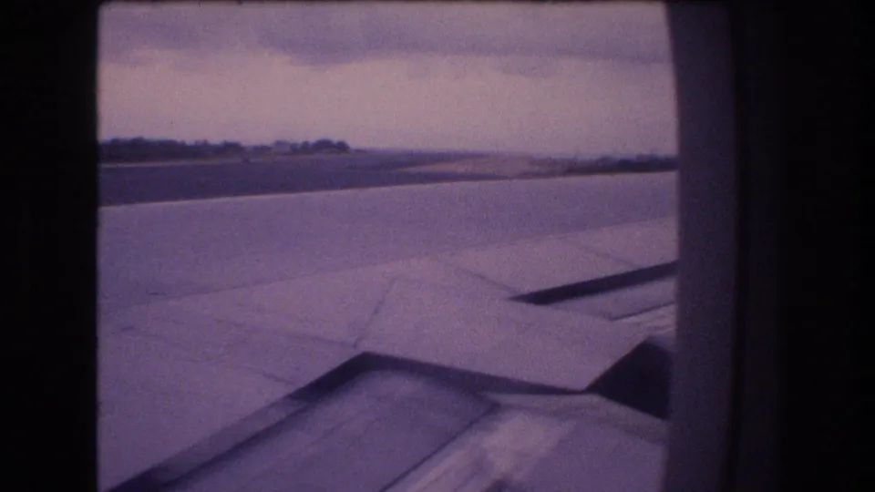 1977 Honolulu airplane-window frame with the wing spanning the foreground, runway pavement below, and a low tree-lined horizon under an overcast sky.