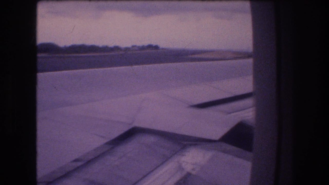 1977 Honolulu, Oahu 8mm home-movie frame showing an aircraft wing and runway area from a passenger window, with distant trees and a muted purple film tint.