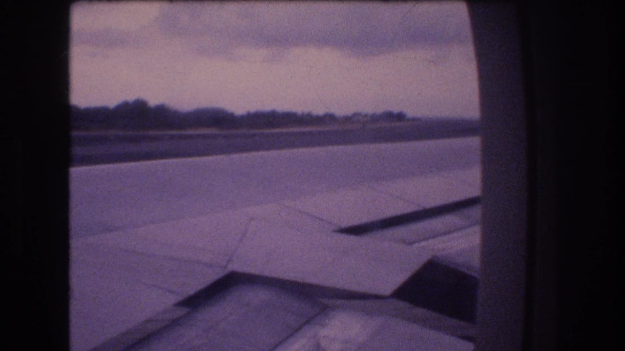 1977 Honolulu, Oahu passenger-window view of an aircraft wing over the runway or taxiway, with a low tree line and soft overcast light in aged 8mm color.