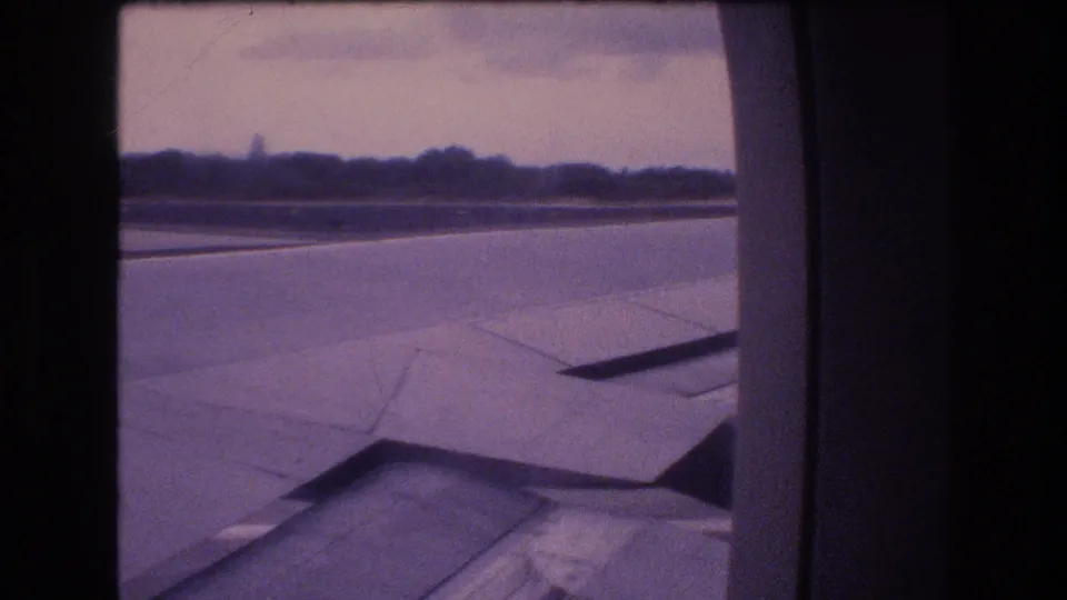 1977 Honolulu, Oahu home-movie frame from a passenger seat showing the aircraft wing, runway area, distant horizon, and heavy purple color fade from the 8mm scan.