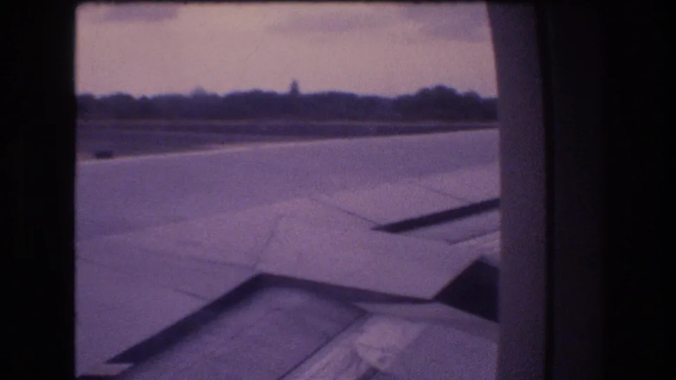 1977 Honolulu, Oahu 8mm frame showing the aircraft wing during takeoff or landing, with runway pavement, a distant tree line, cloudy sky, and visible home-movie grain.