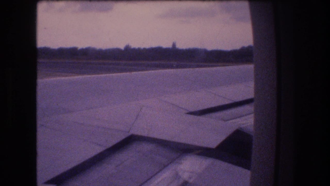 1977 Honolulu, Oahu 8mm frame showing an aircraft wing and flap surface over runway pavement with a distant treeline under a hazy sky.