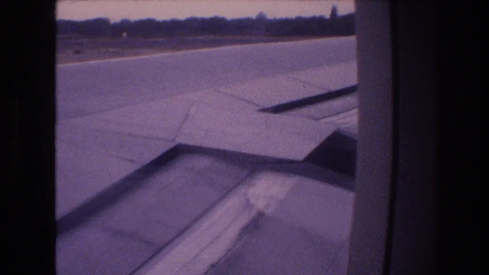 1977 Honolulu, Oahu home-movie frame showing a passenger-seat view across the aircraft wing toward runway pavement and a low treeline.