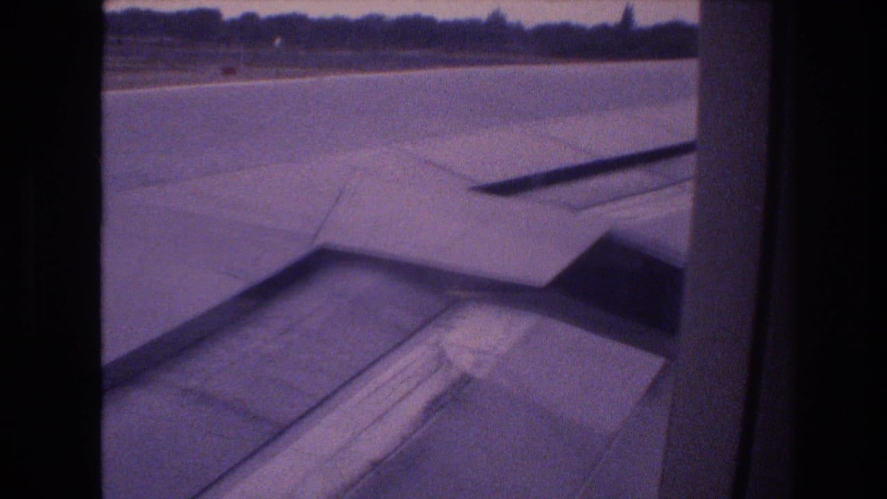 1977 Honolulu home-movie frame showing an airplane wing and flap area viewed from a passenger window while the aircraft is on airport pavement.