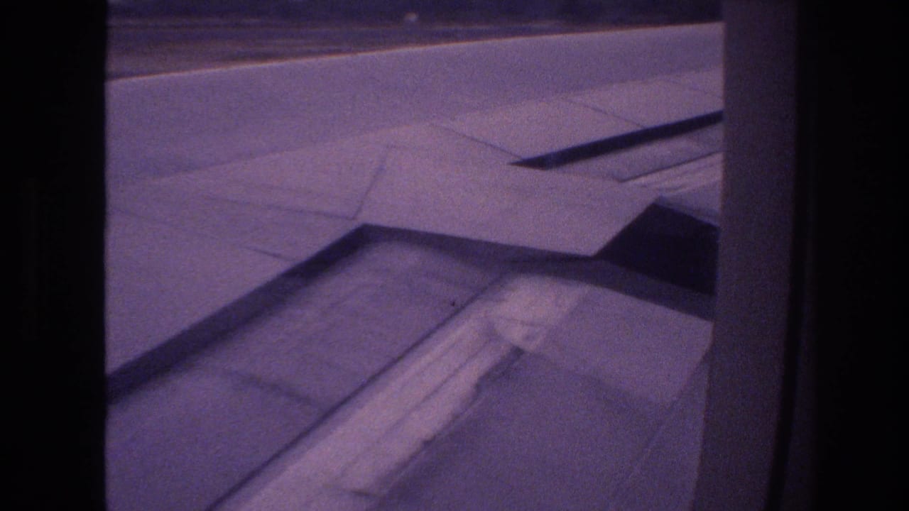 Passenger-window view in Honolulu showing the airplane wing, flap fairing, and runway surface in grainy 8mm footage from 1977.