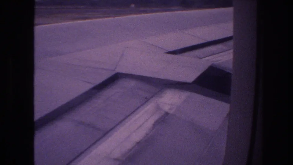 Passenger-window view in Honolulu showing the airplane wing, flap fairing, and runway surface in grainy 8mm footage from 1977.