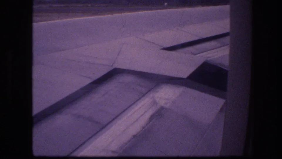 Grainy 1977 Honolulu aircraft-window frame with the wing and flap structure visible as the plane appears to move along the runway or taxiway.