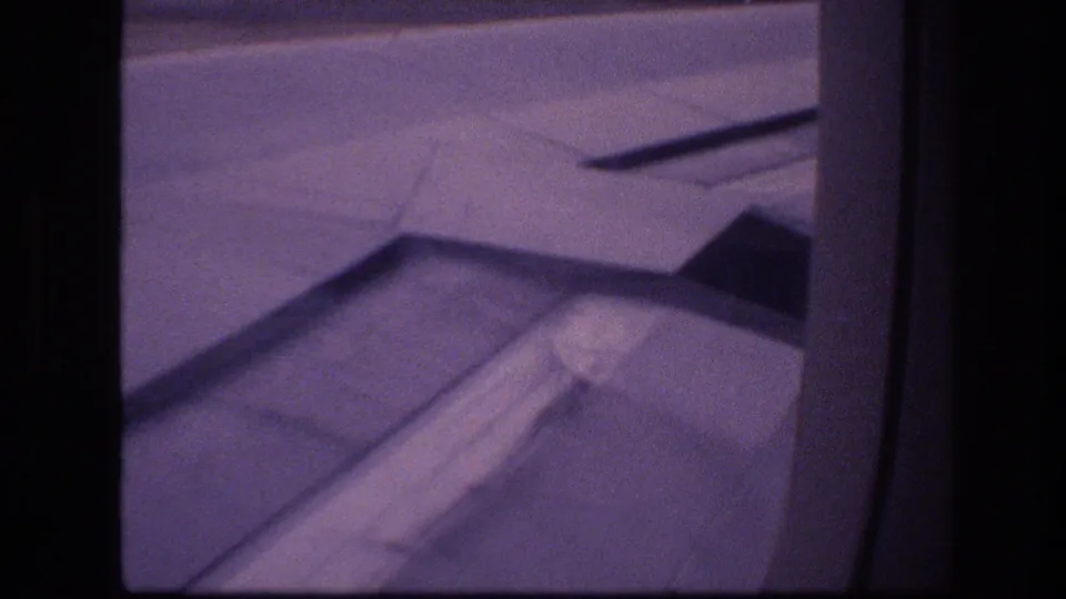Final 1977 Honolulu 8mm frame showing the aircraft wing and flap fairing from inside the plane, with runway pavement and dark window framing visible.