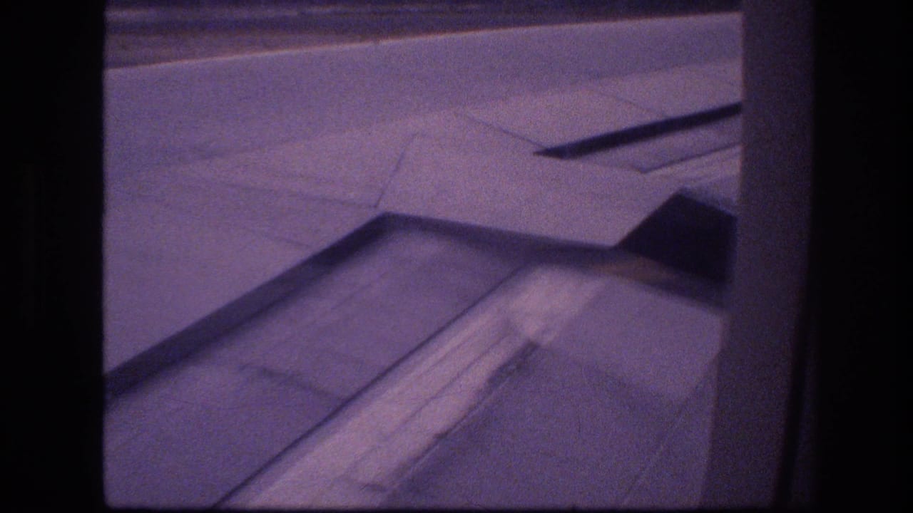 1977 Honolulu-area travel footage showing an airplane wing and flap panels through a passenger window, with runway pavement and a low horizon beyond.