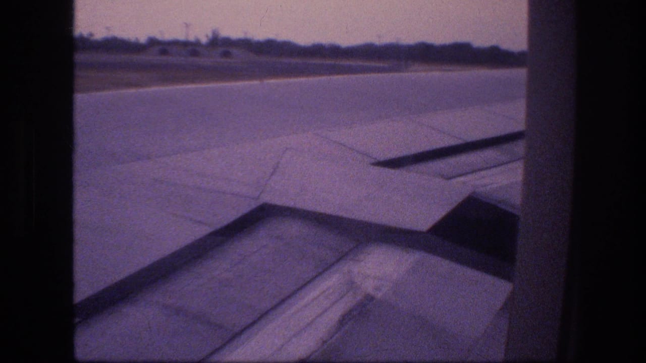 1977 Honolulu, Oahu home-movie frame showing an aircraft wing and runway through a passenger window during takeoff.