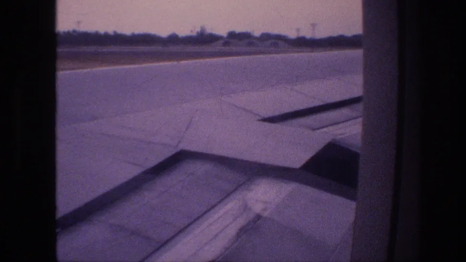 Home-movie frame from a 1977 Honolulu takeoff showing a slightly shifted cabin-window view of the wing, runway, and distant landscape.