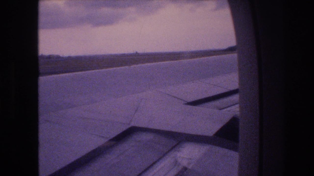 1977 8mm home-movie frame from a flight associated with Honolulu, Oahu, showing an airplane wing through a passenger window with clouds and horizon beyond.