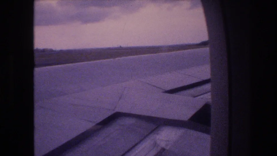 1977 8mm home-movie frame from a flight associated with Honolulu, Oahu, showing an airplane wing through a passenger window with clouds and horizon beyond.