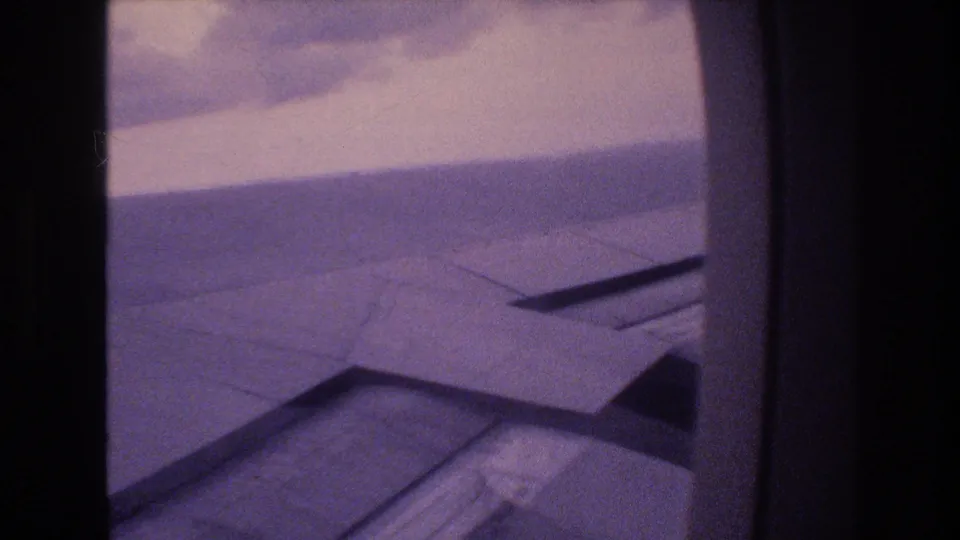 The 8mm frame presents a slightly tilted passenger-window view of the airplane wing under a cloudy sky, with aged purple film coloration visible across the scene.