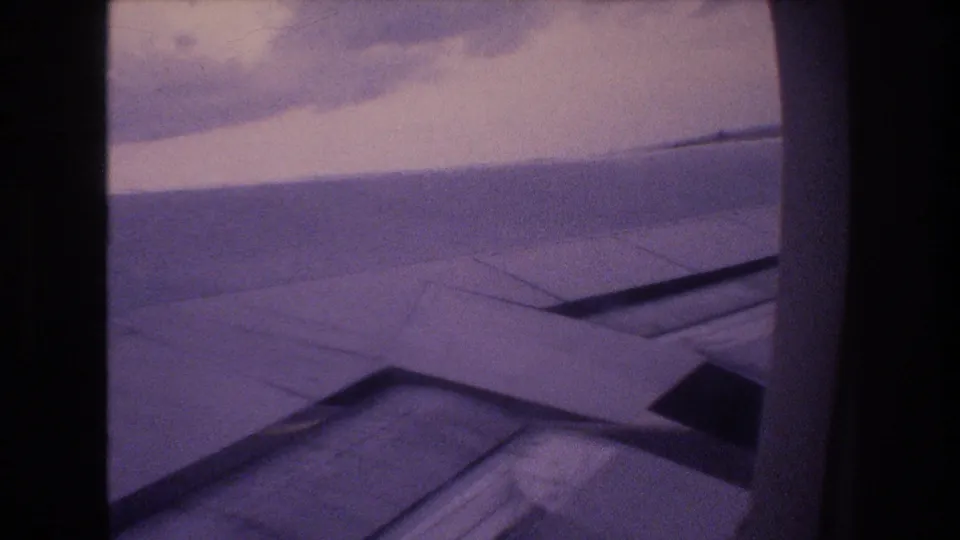 Final 1977 8mm frame shows the airplane wing through a passenger window during flight, with cloudy sky, horizon, film grain, and dark frame edges.