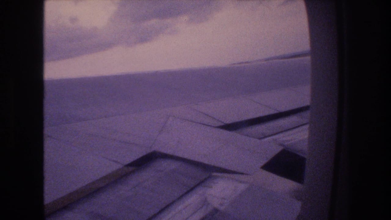 1977 Honolulu-bound home-movie frame showing an aircraft wing through a rounded passenger window during an overcast landing approach.