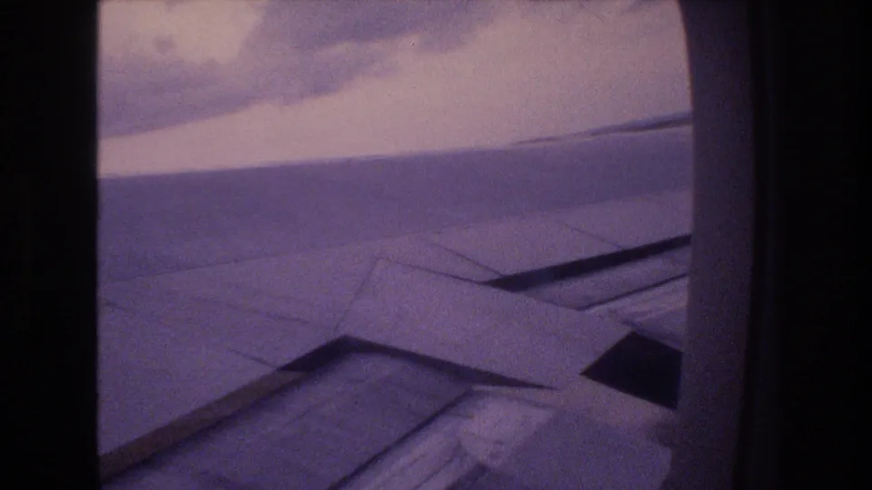 1977 Honolulu-bound home-movie frame showing an aircraft wing through a rounded passenger window during an overcast landing approach.