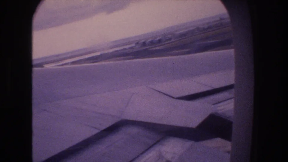 Passenger-window view from 1977 travel footage with the aircraft wing low in frame and a slanted cloud horizon above.
