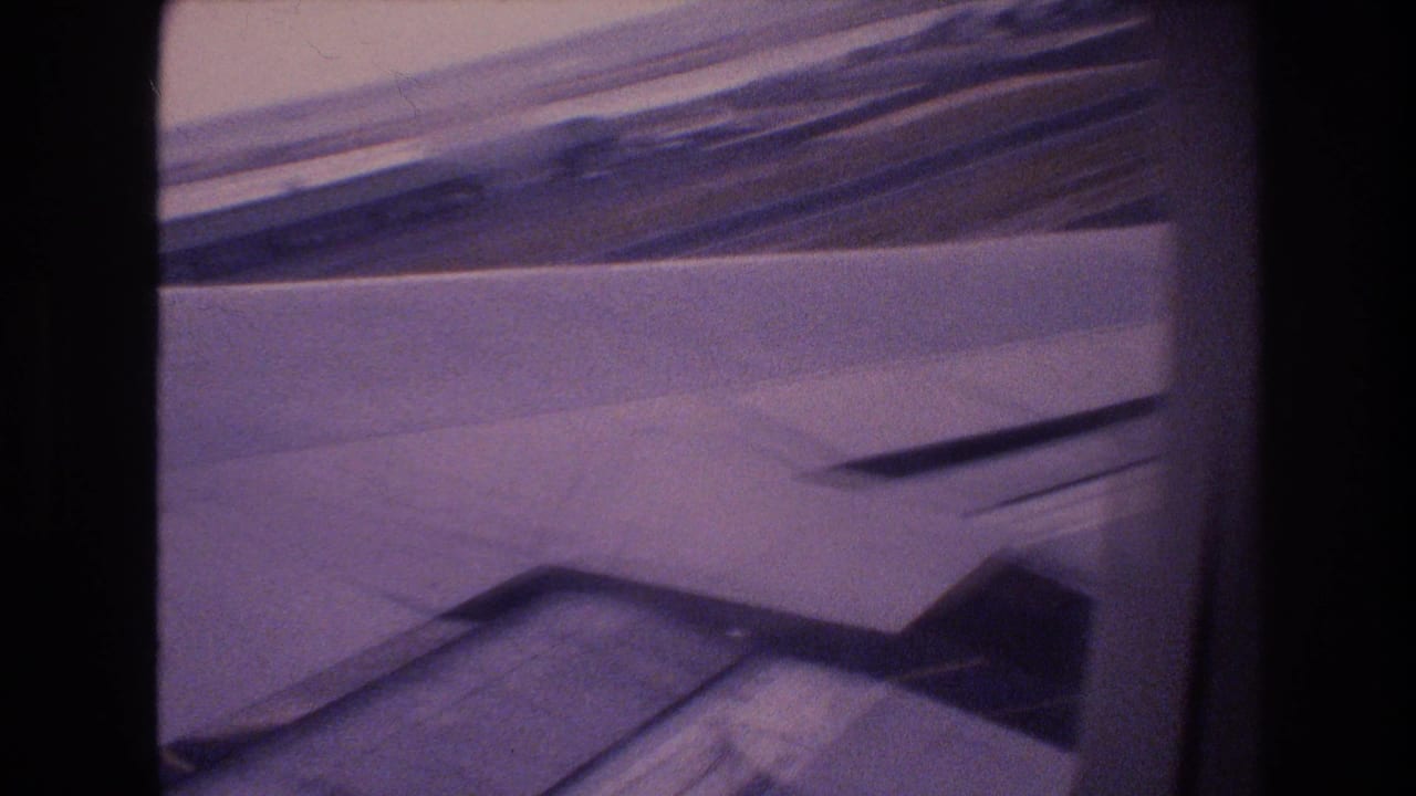 1977 Honolulu Oahu 8mm frame showing an airplane wing and flap area through a passenger window with distant coastline or runway surroundings beyond.