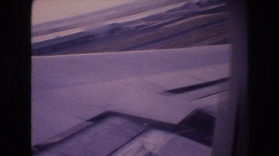1977 Honolulu Oahu 8mm frame showing an airplane wing and flap area through a passenger window with distant coastline or runway surroundings beyond.