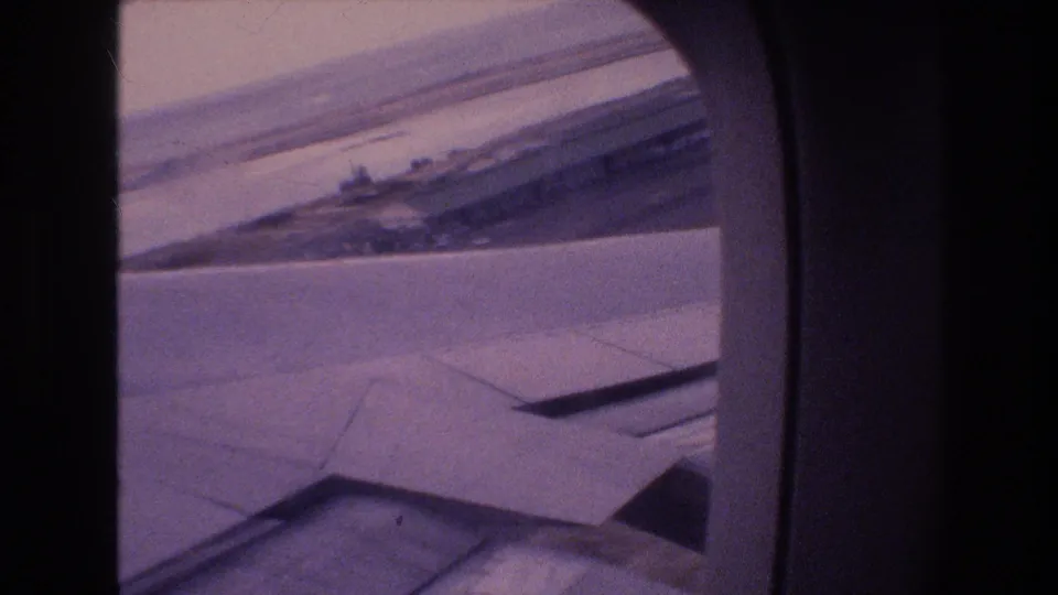 1977 Honolulu Oahu 8mm frame continuing the passenger-window view as the airplane wing crosses a runway or coastal airport landscape.