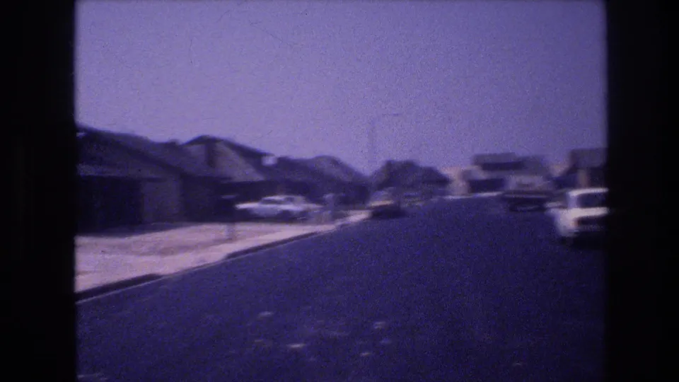 1977 Las Vegas 8mm frame showing a paved suburban street lined with low houses, sidewalks, driveways, and parked cars under a bright sky.