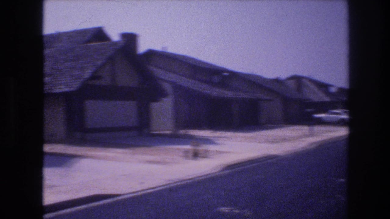1977 Las Vegas street-level frame with a low residential building, sidewalk, curb, parked cars, and a broad paved road continuing through the neighborhood.