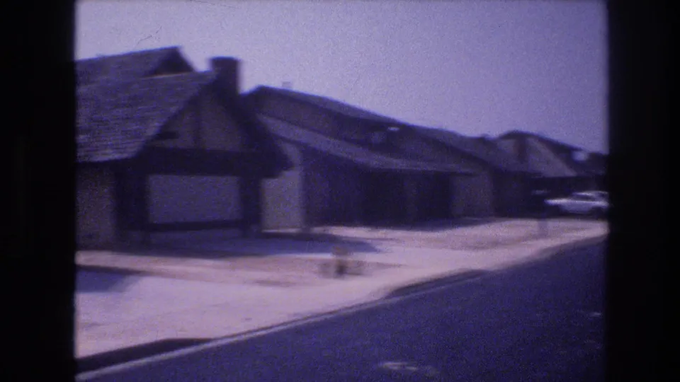 1977 Las Vegas street-level frame with a low residential building, sidewalk, curb, parked cars, and a broad paved road continuing through the neighborhood.
