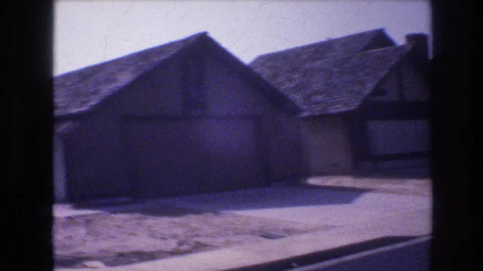 1977 Las Vegas 8mm frame showing closer residential exteriors with sloped roofs, a driveway, sidewalk, curb, and the edge of the street.