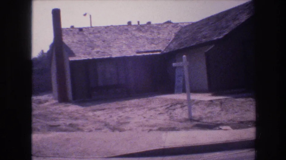 1977 Las Vegas 8mm frame showing a low residential exterior, shingled roof, sidewalk, signpost, bare yard area, and street curb.