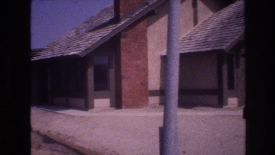 1977 Las Vegas home-movie frame showing two adjacent suburban house exteriors, a pale sidewalk, dusty ground, and strong sun under a clear sky.