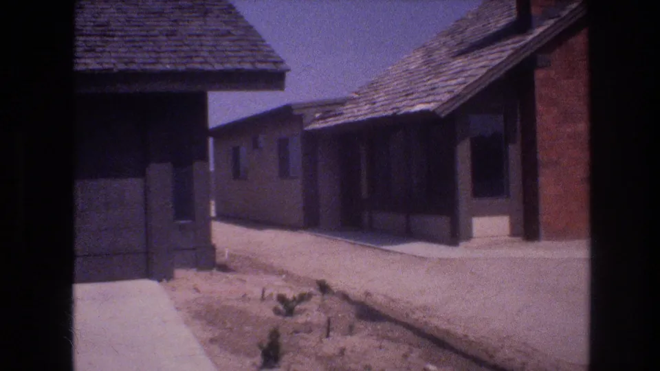 1977 Las Vegas home-movie frame showing adjoining house exteriors, roof shingles, a sidewalk, dusty bare ground, and sparse desert planting.