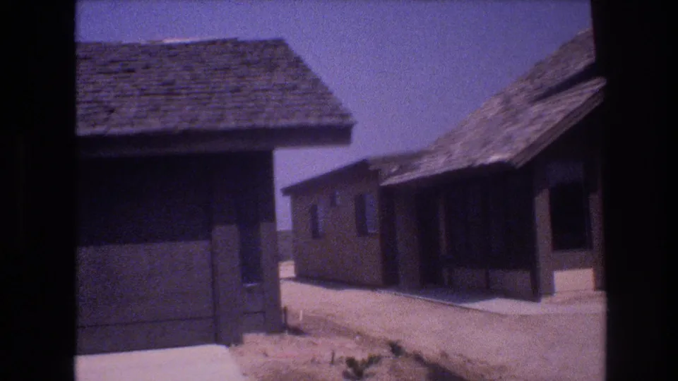 1977 Las Vegas 8mm frame with a shadowed suburban house exterior, adjacent roofline, pale walkway, dusty ground, and clear desert sky.