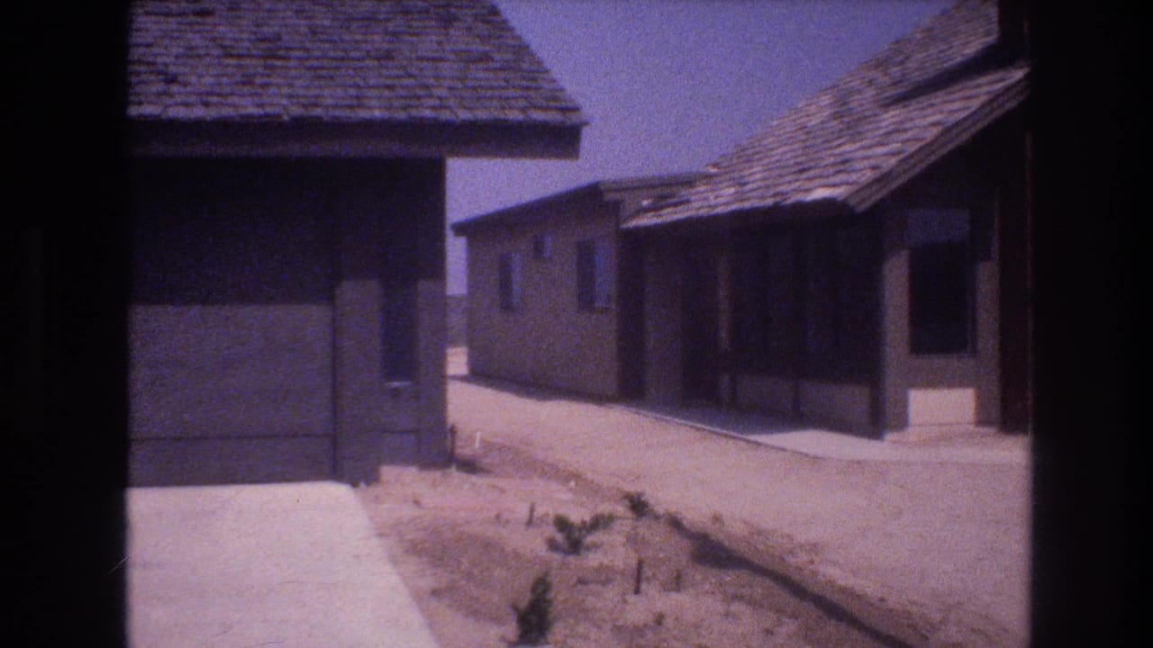 1977 Las Vegas frame showing a quiet row of low stucco houses with garage openings, roof shingles, concrete edges, and unlandscaped desert ground.
