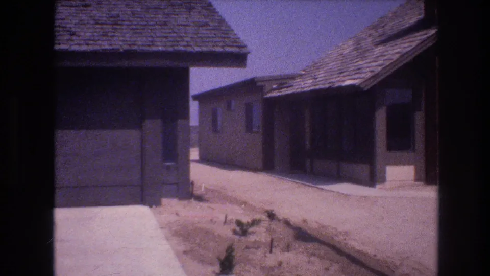1977 Las Vegas frame showing a quiet row of low stucco houses with garage openings, roof shingles, concrete edges, and unlandscaped desert ground.