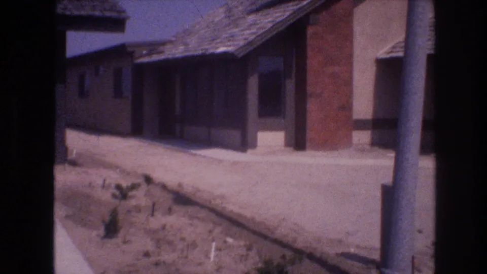 1977 Las Vegas frame showing a stucco house with an attached garage, shingled roof, dirt road, bare lot, small desert plants, and a utility pole at the edge.