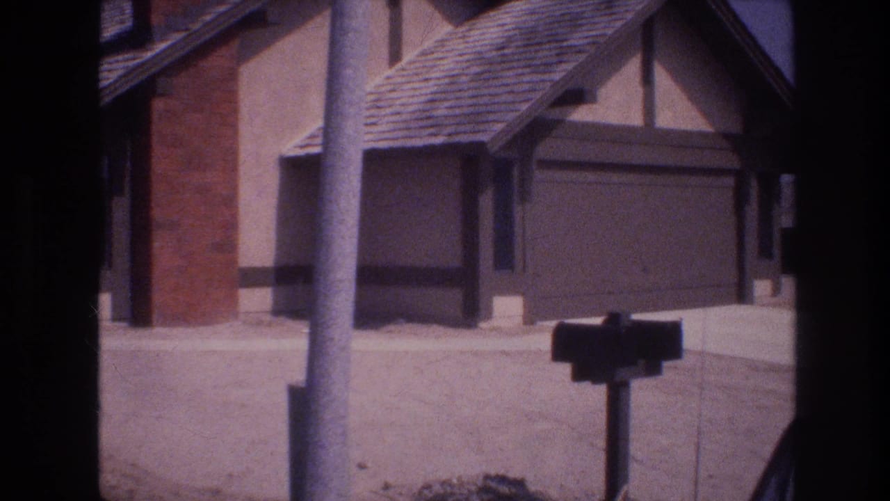 1977 Las Vegas 8mm still of a suburban home exterior, with a utility pole crossing the frame, a dark mailbox, brick chimney, and attached garage.